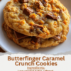 A stack of thick homemade Butterfinger caramel cookies with golden, crispy edges and soft, chewy centers. The cookies are packed with crushed Butterfinger candy pieces and gooey caramel bits, with melted caramel stretching from a broken cookie. Served on a rustic wooden board with scattered candy pieces in warm natural light, creating a cozy bakery-style dessert presentation.