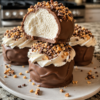 A tray of homemade frozen Cool Whip truffles coated in glossy milk chocolate, with a few topped with crushed toffee bits, chopped nuts, and mini chocolate chips. One truffle is cut open to reveal the light, fluffy, creamy center. Served on parchment paper in warm natural light on a rustic wooden table for an elegant no-bake dessert presentation.