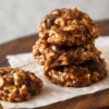 A stack of homemade no-bake oatmeal cookies made with chewy quick oats, rich caramel, melted butterscotch chips, and chocolate chips, arranged on parchment paper. The cookies have a glossy golden-brown finish with visible chocolate and toffee pieces, photographed in warm natural light on a rustic wooden table for a cozy homemade dessert presentation.