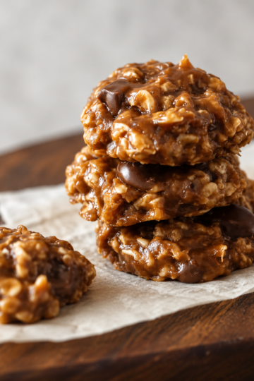 A stack of homemade no-bake oatmeal cookies made with chewy quick oats, rich caramel, melted butterscotch chips, and chocolate chips, arranged on parchment paper. The cookies have a glossy golden-brown finish with visible chocolate and toffee pieces, photographed in warm natural light on a rustic wooden table for a cozy homemade dessert presentation.