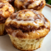 Golden-brown cinnamon roll muffins with soft, tender centers and visible swirls of cinnamon sugar throughout. The muffins are generously drizzled with sweet vanilla glaze that melts into the warm tops, creating a cozy homemade brunch treat.