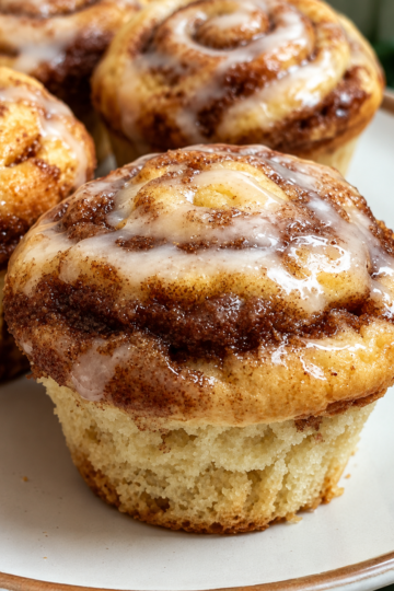 Golden-brown cinnamon roll muffins with soft, tender centers and visible swirls of cinnamon sugar throughout. The muffins are generously drizzled with sweet vanilla glaze that melts into the warm tops, creating a cozy homemade brunch treat.