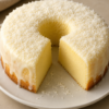 A creamy coconut custard cake baked until lightly golden and coated with sweet shredded coconut flakes. A slice has been removed to reveal the smooth, silky interior with a flan-like texture. Served on a white cake stand with extra toasted coconut scattered around, photographed in warm natural light on a rustic wooden table for an elegant homemade tropical dessert presentation.