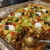 Walking Taco Bake made with seasoned ground beef, chili beans, crunchy corn chips, and melted cheddar cheese baked together in a casserole dish. The golden, bubbly cheese is topped with fresh lettuce, diced tomatoes, black olives, jalapeños, sour cream, and green onions for a colorful, loaded taco-inspired dinner.