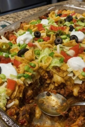 Walking Taco Bake made with seasoned ground beef, chili beans, crunchy corn chips, and melted cheddar cheese baked together in a casserole dish. The golden, bubbly cheese is topped with fresh lettuce, diced tomatoes, black olives, jalapeños, sour cream, and green onions for a colorful, loaded taco-inspired dinner.