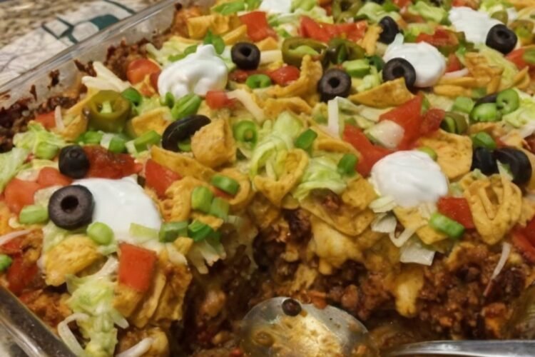 Walking Taco Bake made with seasoned ground beef, chili beans, crunchy corn chips, and melted cheddar cheese baked together in a casserole dish. The golden, bubbly cheese is topped with fresh lettuce, diced tomatoes, black olives, jalapeños, sour cream, and green onions for a colorful, loaded taco-inspired dinner.