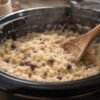 Creamy slow cooker rice pudding made with tender short-grain rice, whole milk, cinnamon, vanilla, and plump raisins. Served warm in a bowl with a light sprinkle of cinnamon on top, showcasing its thick, custardy texture and classic old-fashioned comfort.