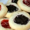 Classic buttery thumbprint cookies with tender, melt-in-your-mouth centers and lightly golden edges, each filled with a glossy jewel of fruit jam. The cookies feature colorful strawberry, raspberry, and blueberry centers and are arranged on a baking sheet as a nostalgic homemade holiday treat.