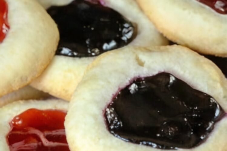 Classic buttery thumbprint cookies with tender, melt-in-your-mouth centers and lightly golden edges, each filled with a glossy jewel of fruit jam. The cookies feature colorful strawberry, raspberry, and blueberry centers and are arranged on a baking sheet as a nostalgic homemade holiday treat.