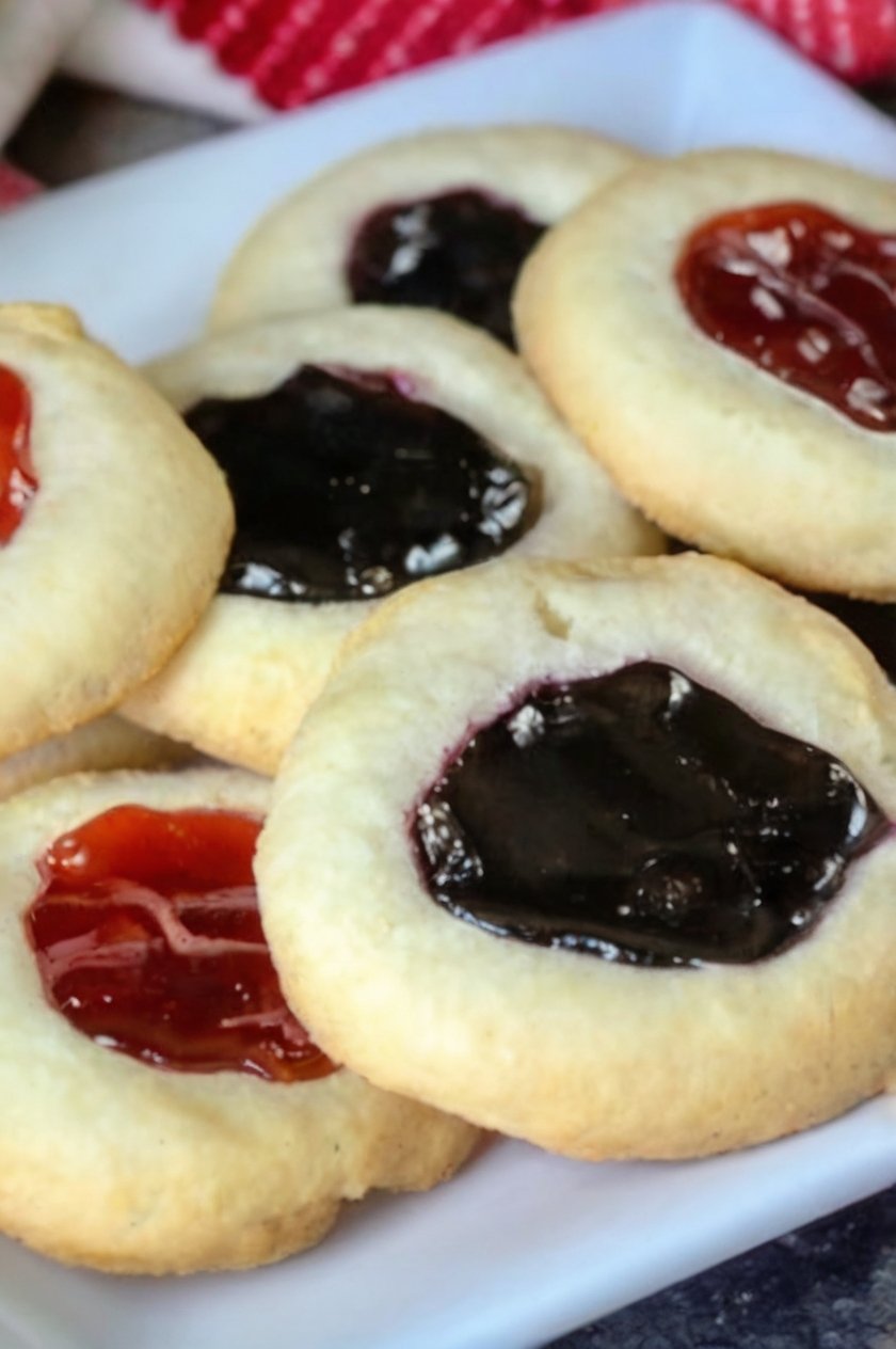 Classic buttery thumbprint cookies with tender, melt-in-your-mouth centers and lightly golden edges, each filled with a glossy jewel of fruit jam. The cookies feature colorful strawberry, raspberry, and blueberry centers and are arranged on a baking sheet as a nostalgic homemade holiday treat.
