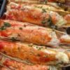 A close-up platter of baked king crab legs coated in rich garlic butter and Creole seasoning, garnished with fresh herbs and served with lemon wedges and a small dish of melted butter for dipping.
