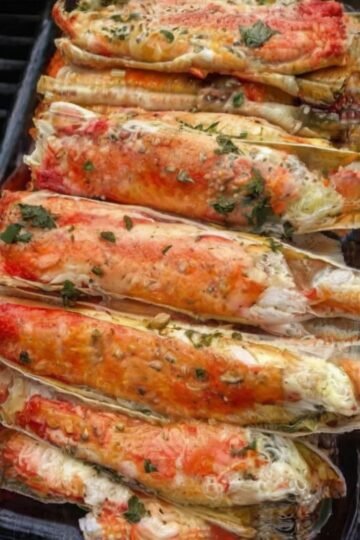 A close-up platter of baked king crab legs coated in rich garlic butter and Creole seasoning, garnished with fresh herbs and served with lemon wedges and a small dish of melted butter for dipping.
