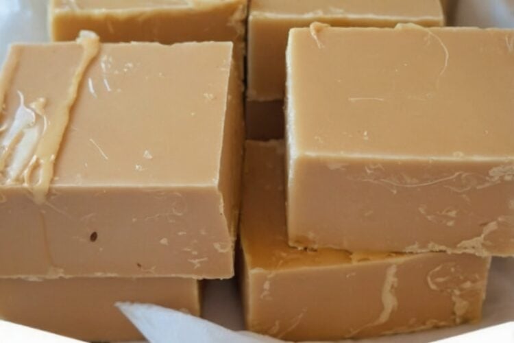 A stack of old-fashioned 3-ingredient peanut butter fudge cut into neat squares with a smooth, creamy texture and a light fudge crumble. The rich peanut butter fudge is arranged on parchment paper atop a rustic wooden board, with a few pieces broken open to show the soft interior. Photographed in warm natural light for a cozy homemade holiday candy presentation.