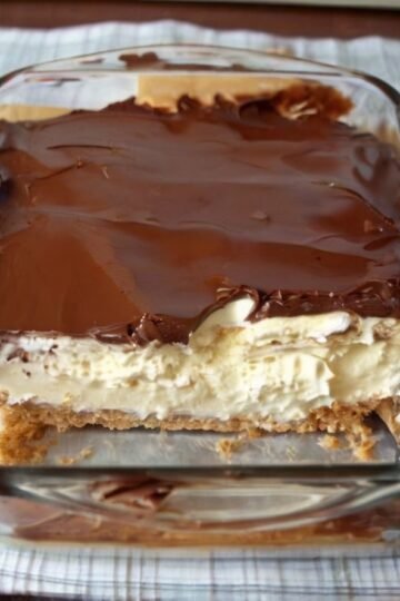A close-up of a no-bake chocolate éclair cake in a glass baking dish, featuring layers of soft graham crackers, fluffy vanilla cream filling, and a thick, glossy chocolate fudge topping spread across the surface.
