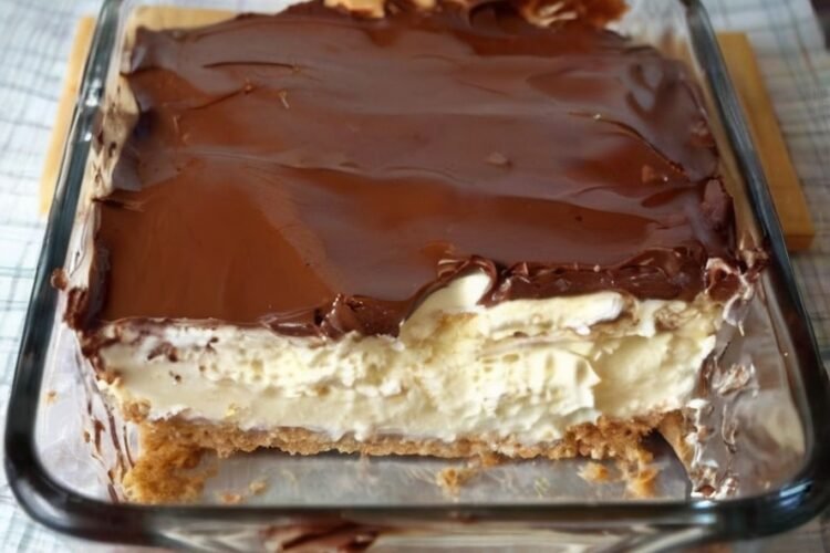 A close-up of a no-bake chocolate éclair cake in a glass baking dish, featuring layers of soft graham crackers, fluffy vanilla cream filling, and a thick, glossy chocolate fudge topping spread across the surface.