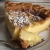 Slice of old-fashioned custard pie with a golden caramelized top, creamy vanilla custard center, and powdered sugar dusting, served on a white plate.