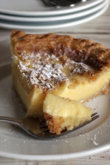 Slice of old-fashioned custard pie with a golden caramelized top, creamy vanilla custard center, and powdered sugar dusting, served on a white plate.
