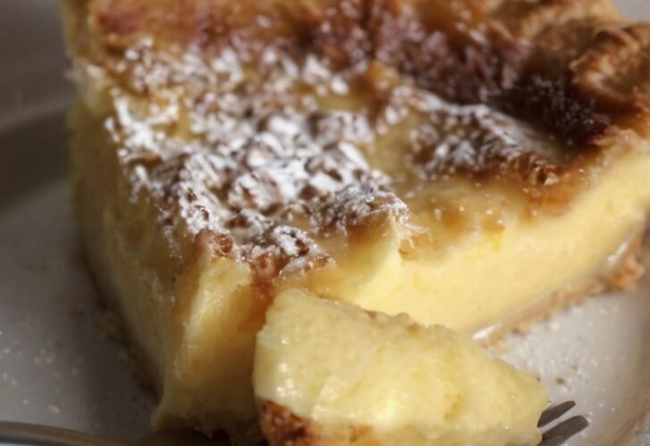 Slice of old-fashioned custard pie with a golden caramelized top, creamy vanilla custard center, and powdered sugar dusting, served on a white plate.