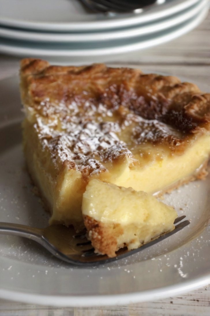 Slice of old-fashioned custard pie with a golden caramelized top, creamy vanilla custard center, and powdered sugar dusting, served on a white plate.