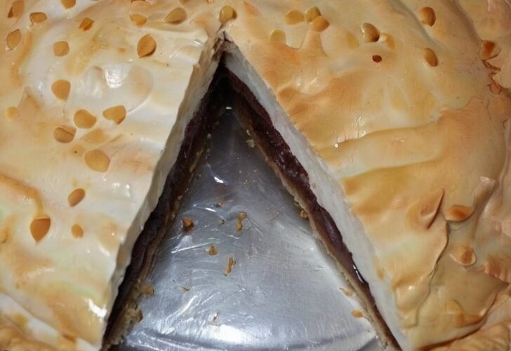 A homemade chocolate meringue pie in a foil pan, topped with a thick layer of golden-brown meringue and caramelized droplets, with a slice removed to reveal the rich chocolate filling and crust underneath.