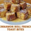 A plate of golden-brown cinnamon roll French toast bites stacked high, with soft cinnamon swirls inside, dusted with powdered sugar and generously drizzled with creamy icing and maple syrup.