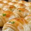 Freshly baked golden crescent-shaped dinner rolls with flaky layers, brushed with butter and garnished with fresh parsley, arranged on a baking tray.