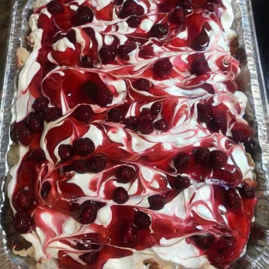 A rectangular tray of Heaven on Earth Cake layered with fluffy cake, creamy white pudding, whipped topping, and swirls of glossy cherry pie filling across the top.