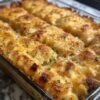 A close-up of a cheesy hashbrown casserole in a rectangular baking dish, with tender shredded potatoes covered in melted golden cheddar cheese and a crispy browned topping. Small flecks of fresh parsley garnish the bubbly, golden surface.