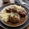Salisbury steak patties topped with mushroom gravy, served with creamy mashed potatoes on a dark plate.