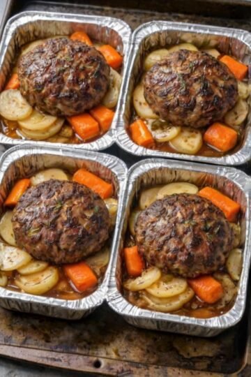 Four individual meatloaf portions baked in aluminum trays with sliced potatoes and carrots, topped with a savory brown glaze.