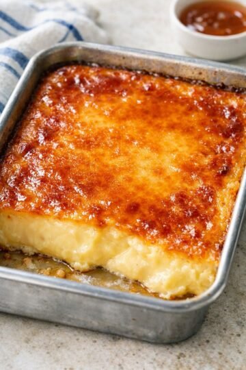 A rich baked custard dessert with a smooth, creamy golden center and a glossy caramelized top, baked in a rectangular metal pan and sliced into thick squares.
