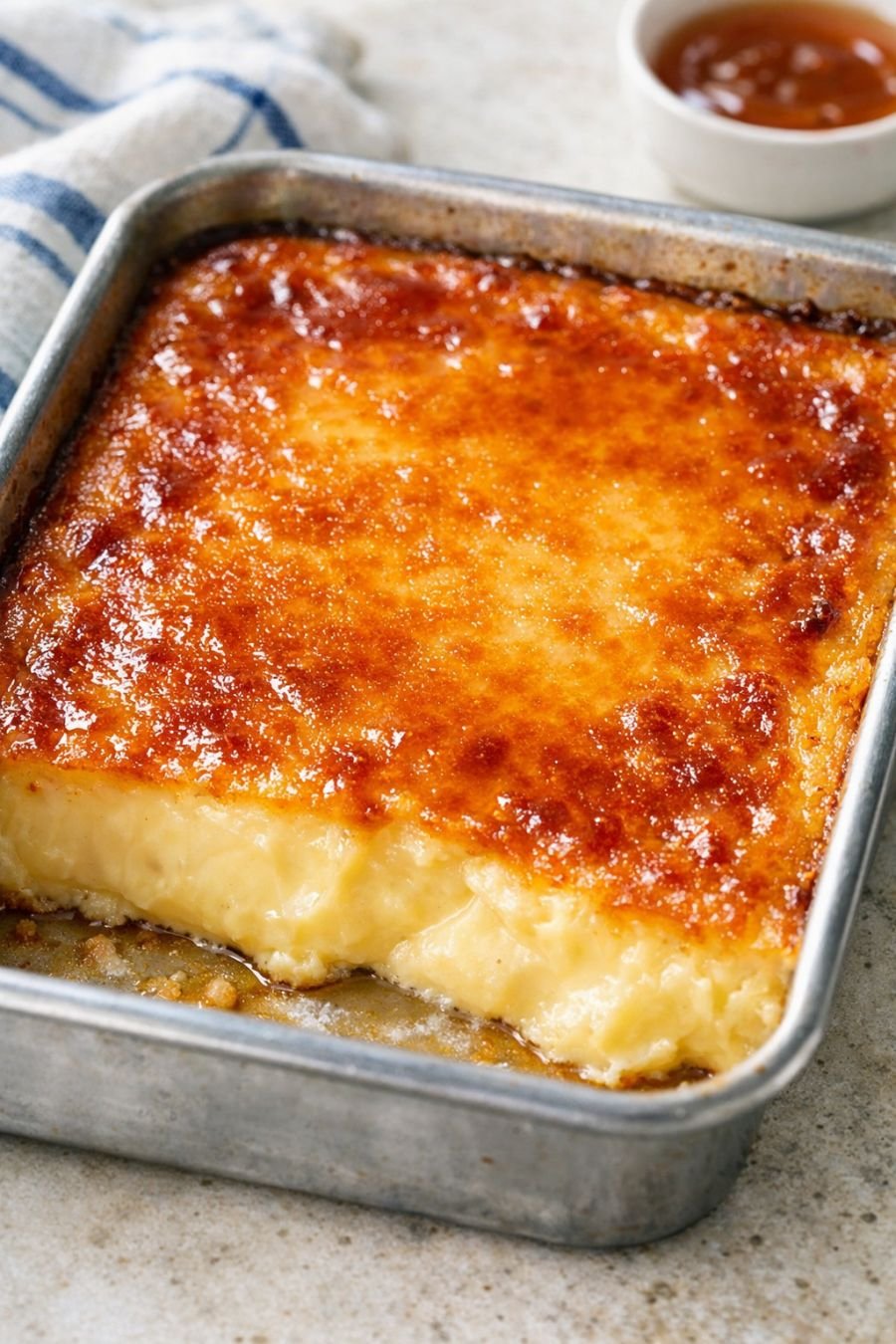 A rich baked custard dessert with a smooth, creamy golden center and a glossy caramelized top, baked in a rectangular metal pan and sliced into thick squares.