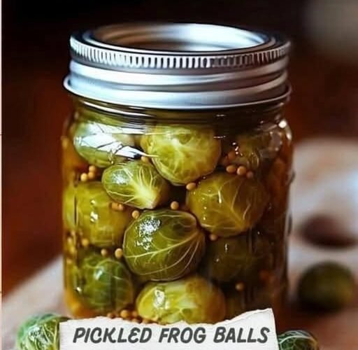 A jar of pickled Brussels sprouts, packed in a tangy-looking brine with mustard seeds and labeled “Pickled Frog Balls,” with a few fresh Brussels sprouts beside the jar. delish.com