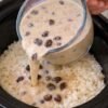 A creamy rice dish in a slow cooker, topped with a rich, velvety sauce studded with plump raisins, being poured over fluffy white rice.