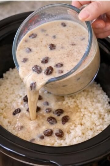 A creamy rice dish in a slow cooker, topped with a rich, velvety sauce studded with plump raisins, being poured over fluffy white rice.
