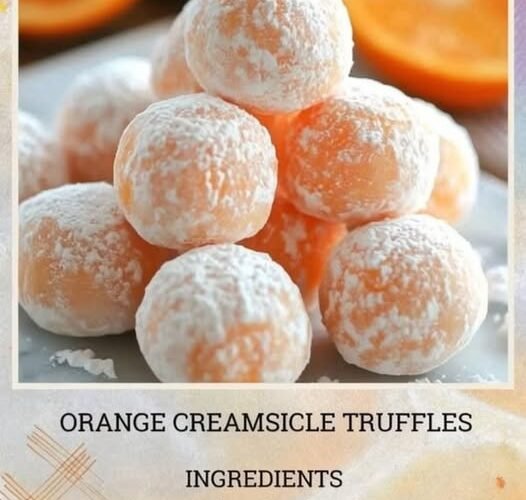 A stack of soft orange creamsicle truffles coated in powdered sugar, with a delicate citrus-orange center and fresh orange slices blurred in the background.