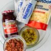 A close-up of ingredients for a sweet-and-savory appetizer, featuring cream cheese, shredded sharp cheddar, raspberry preserves, chopped jalapeños, and crispy bacon pieces arranged on a plate.