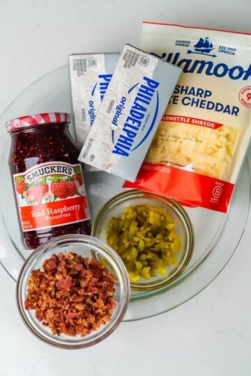 A close-up of ingredients for a sweet-and-savory appetizer, featuring cream cheese, shredded sharp cheddar, raspberry preserves, chopped jalapeños, and crispy bacon pieces arranged on a plate.