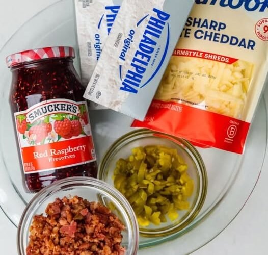 A close-up of ingredients for a sweet-and-savory appetizer, featuring cream cheese, shredded sharp cheddar, raspberry preserves, chopped jalapeños, and crispy bacon pieces arranged on a plate.