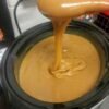 A rich, silky caramel sauce with a glossy golden-brown finish, slowly cascading from a spoon into a black slow cooker and creating smooth swirls on the surface.
