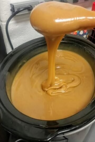 A rich, silky caramel sauce with a glossy golden-brown finish, slowly cascading from a spoon into a black slow cooker and creating smooth swirls on the surface.
