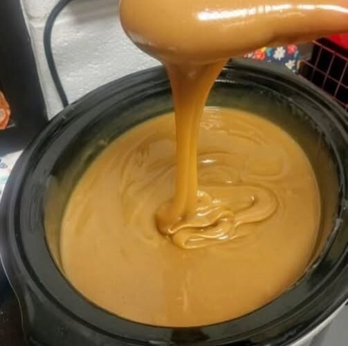 A rich, silky caramel sauce with a glossy golden-brown finish, slowly cascading from a spoon into a black slow cooker and creating smooth swirls on the surface.