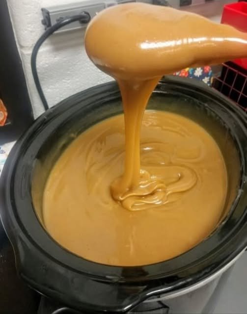 A rich, silky caramel sauce with a glossy golden-brown finish, slowly cascading from a spoon into a black slow cooker and creating smooth swirls on the surface.