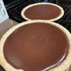Two homemade chocolate pies with smooth, glossy chocolate filling nestled in golden, crimped pastry crusts, cooling on a wire rack.