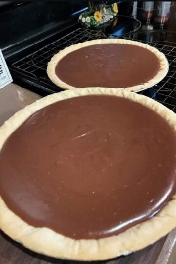 Two homemade chocolate pies with smooth, glossy chocolate filling nestled in golden, crimped pastry crusts, cooling on a wire rack.