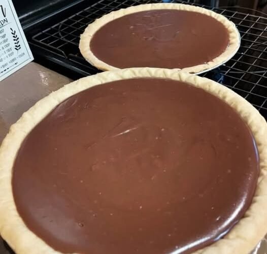 Two homemade chocolate pies with smooth, glossy chocolate filling nestled in golden, crimped pastry crusts, cooling on a wire rack.