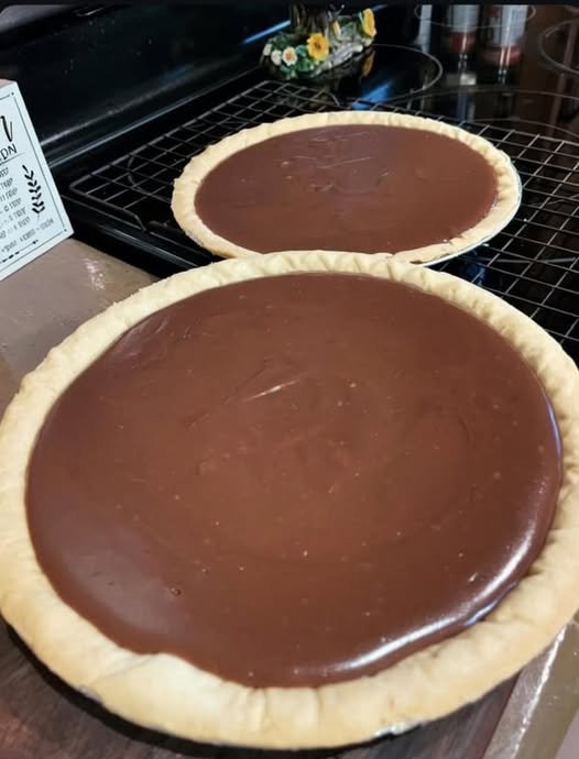 Two homemade chocolate pies with smooth, glossy chocolate filling nestled in golden, crimped pastry crusts, cooling on a wire rack.