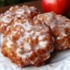 A plate of golden-brown apple fritters coated in a glossy sweet glaze, with tender apple pieces throughout and fresh red apples in the background.