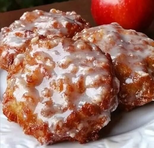 A plate of golden-brown apple fritters coated in a glossy sweet glaze, with tender apple pieces throughout and fresh red apples in the background.