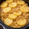 A rustic slow cooker potato and onion bake with tender golden potato slices layered over sweet caramelized onions, lightly coated in melted butter and seasoning, served hot in a black slow cooker.